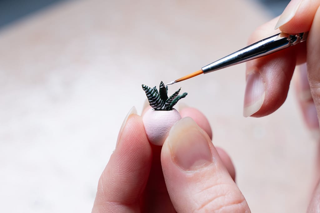 A fine paint brush applying white paint to a miniature succulent plant in a pot held between four fingers